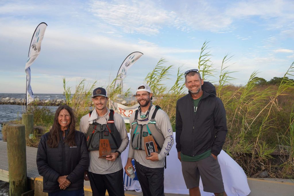 Group Of Paddlers With Lifejackets And Trophies With Dunes And Reeds And The Ocean In The Background